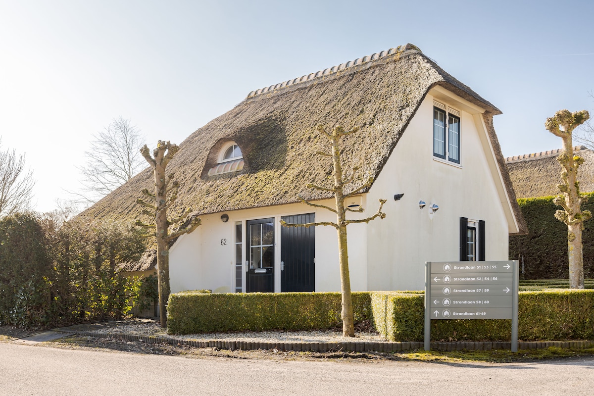 A charming vacation home features a thatched roof and is surrounded by neatly trimmed hedges. The entrance, framed by a pair of trees, leads to an inviting front door. A sign with accommodation information is positioned in front of the property.