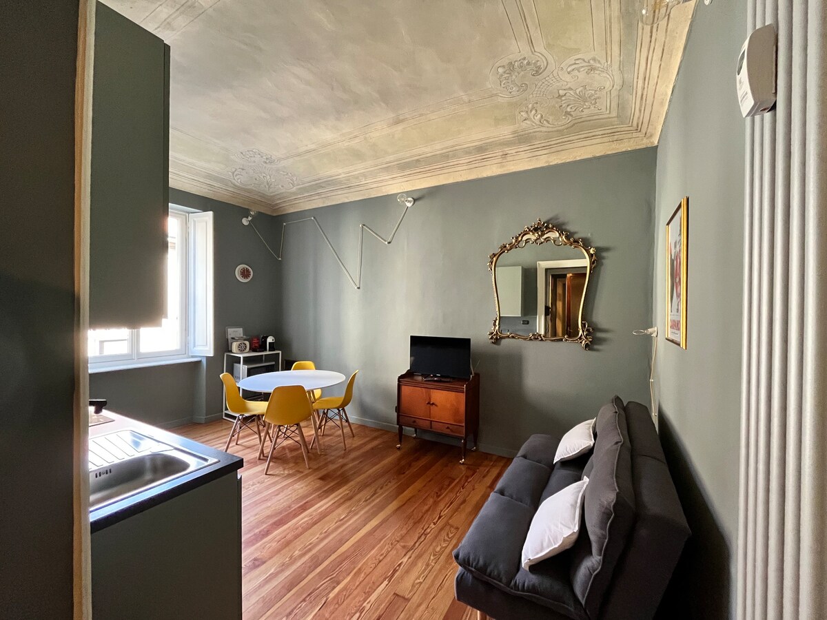A living area features a modern dining table with yellow chairs and a wooden cabinet. A television is positioned against one wall, while a stylish mirror adorns another. Natural light fills the space through a large window, highlighting the intricate ceiling design above.