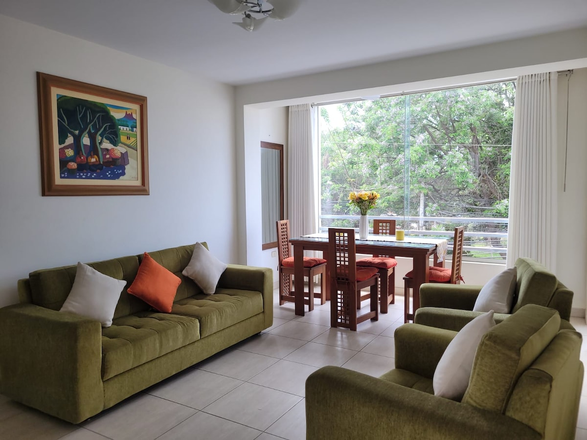 A cozy living area is arranged with two green sofas, accentuated by orange and white pillows. A wooden dining table with matching chairs is set near a large window, allowing natural light to brighten the space. A colorful painting is displayed on the wall.