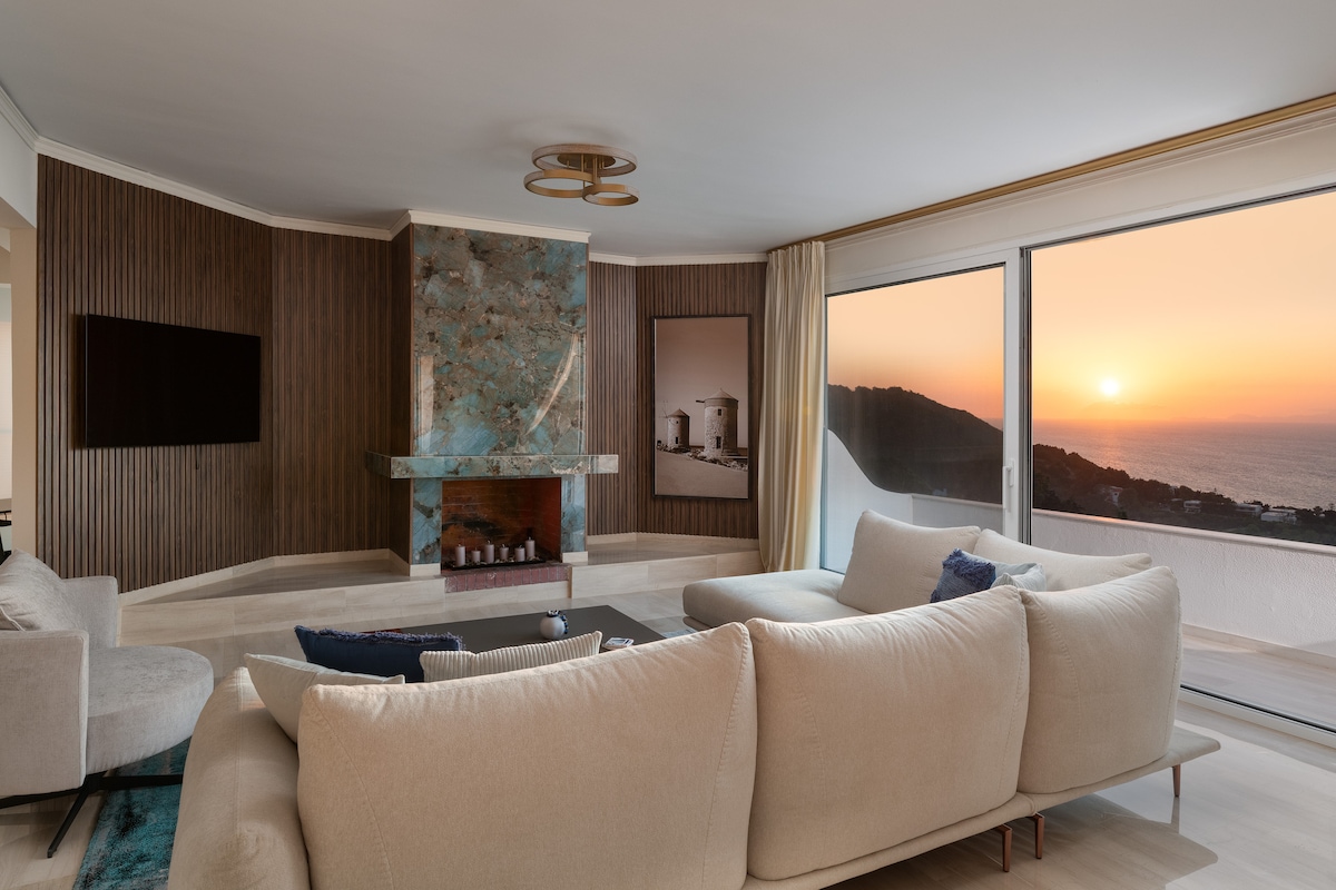 A spacious living room features a modern design with a large sectional sofa positioned around a sleek coffee table. A stone fireplace with blue accents stands as a focal point, while expansive windows frame a stunning sunset view over the Aegean Sea.