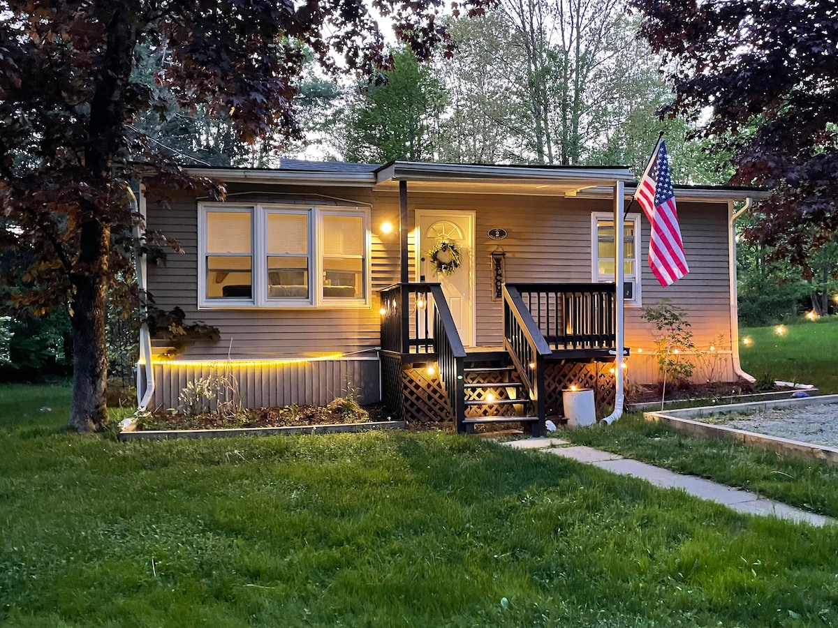 A charming cottage exterior is framed by lush green grass and trees. The inviting front steps lead to a modest porch, adorned with a welcoming wreath. An American flag is displayed prominently, while soft lighting illuminates the house as dusk settles in.
