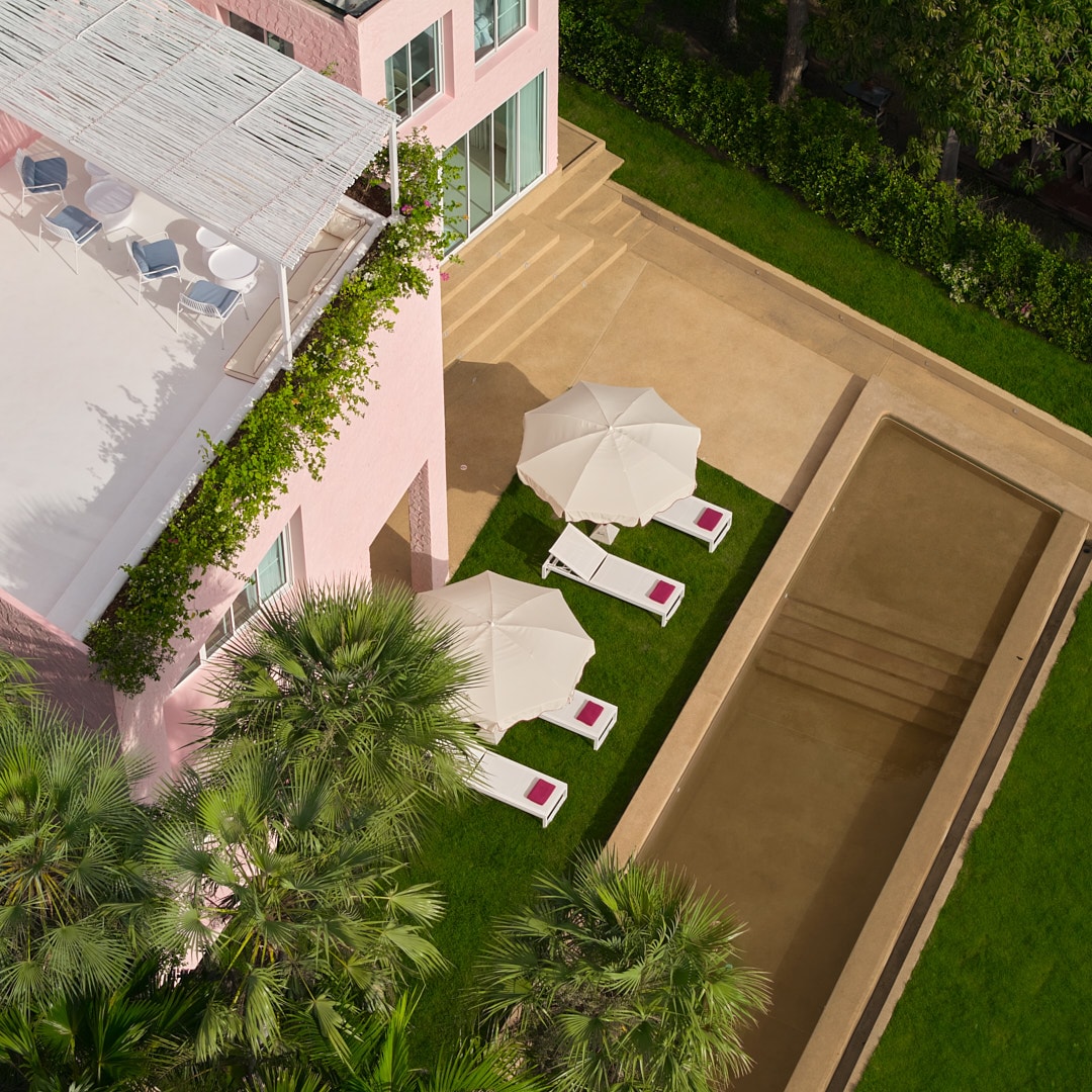 An aerial view captures the property’s outdoor area, featuring a rectangular pool bordered by lush greenery. Sun loungers with cream-colored umbrellas are positioned on the lawn, enhancing the space's relaxation options. The building’s pink exterior contrasts with the natural surroundings.