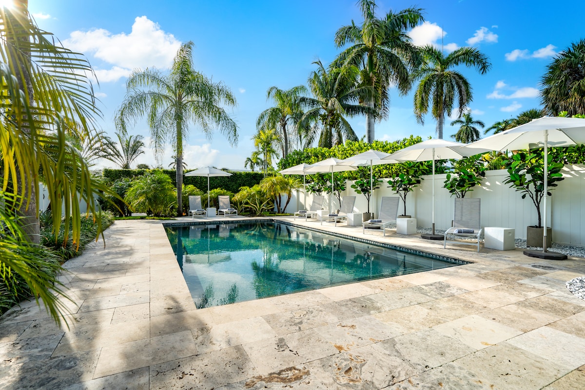 A private saltwater pool is surrounded by travertine stone and lush palm trees. Plush loungers and shaded umbrellas offer relaxation options. The tranquil water reflects the clear blue sky, providing an inviting space for leisure and enjoyment.