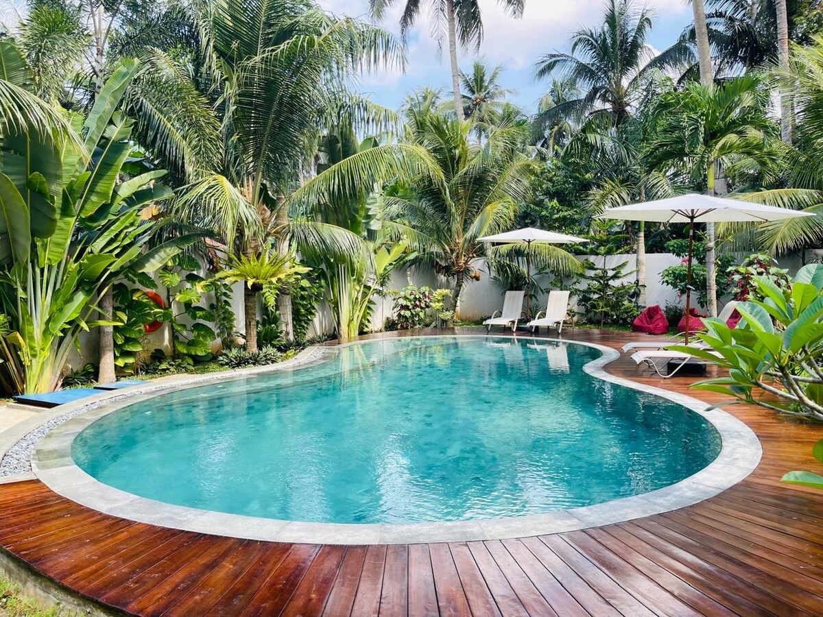 A landscaped outdoor area features a sparkling blue swimming pool surrounded by lush palm trees and vibrant tropical plants. Several sun loungers are positioned near the pool, and two umbrellas provide shade, creating a relaxing environment for guests.