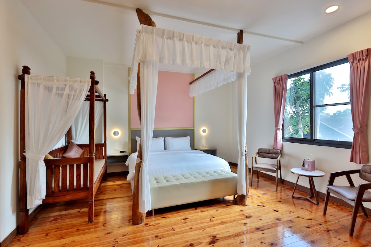 A spacious guest room features a four-poster bed with soft bedding, complemented by an additional wooden cradle. Natural light streams in through a large window adorned with light curtains, enhancing the warm wooden flooring and minimalist decor.