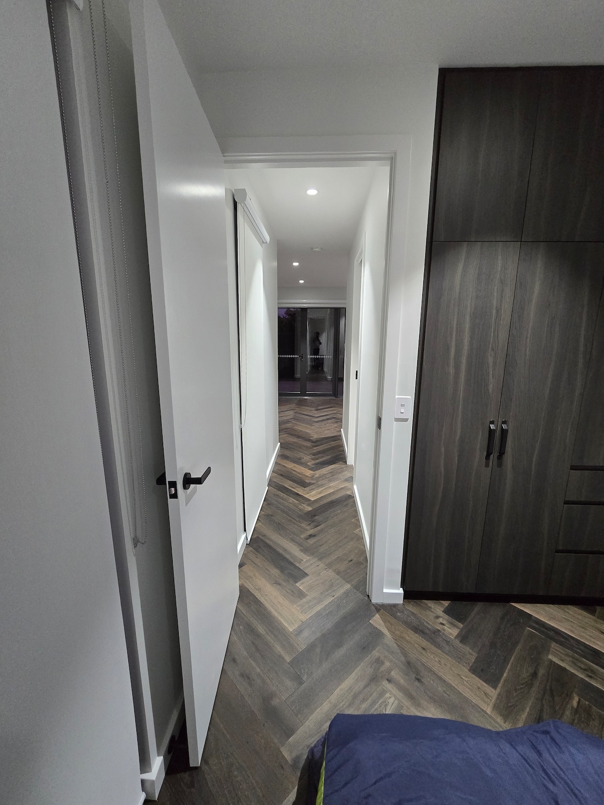A narrow hallway is viewed from the bedroom, showcasing a herringbone patterned wooden floor. White walls and multiple light fixtures enhance the brightness. A door on the left leads to the bedroom, while the hallway extends towards a darker area, creating a sense of depth.