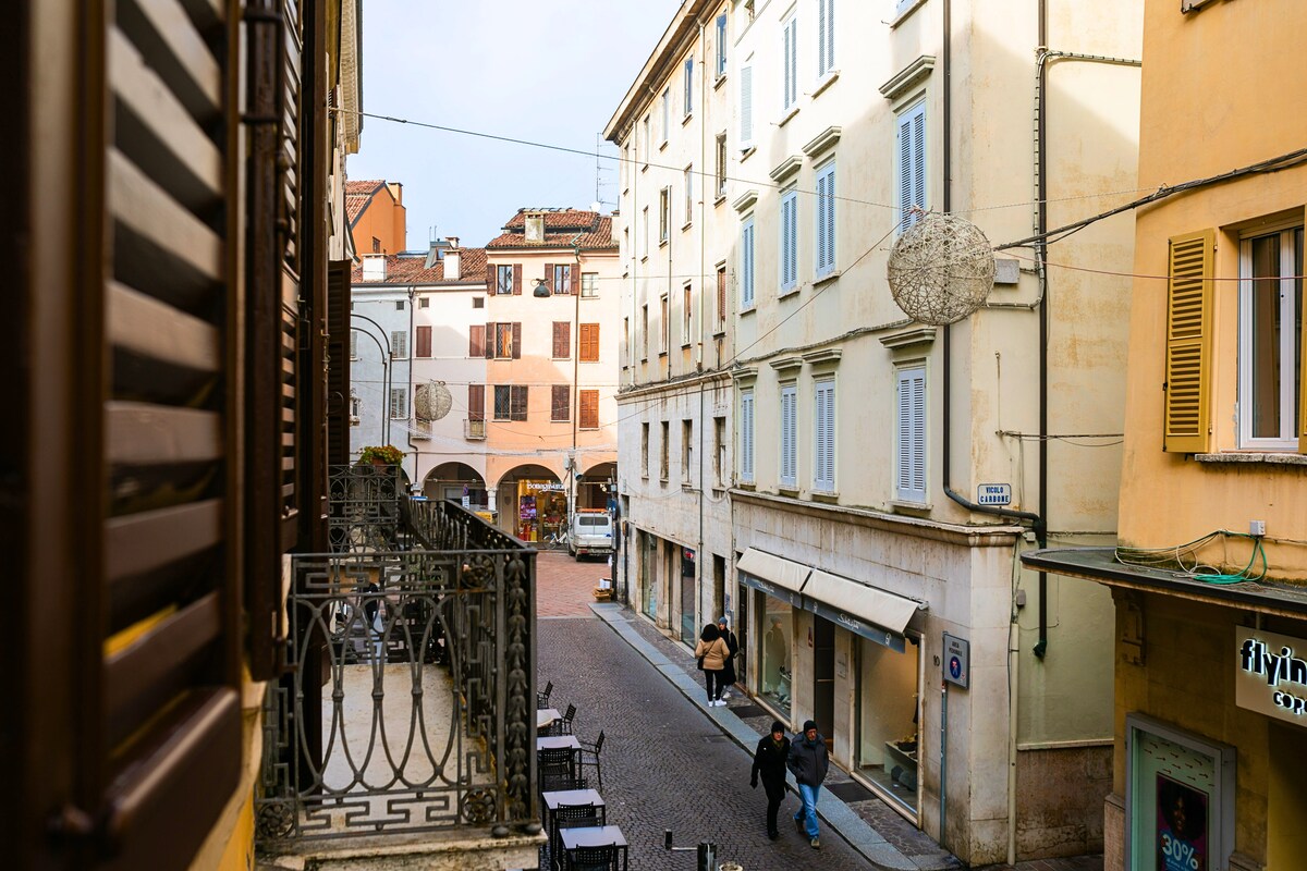 A view from a balcony reveals a charming street in the historic center of Mantova, featuring light-colored buildings and narrow cobblestone paths. Pedestrians are seen walking, with various shops and a cafe present along the street. Natural light enhances the inviting atmosphere.