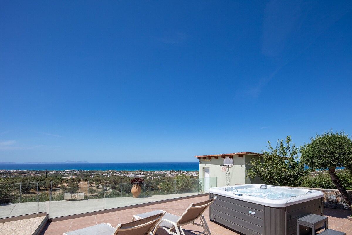 A heated five-seater jacuzzi is positioned on a spacious terrace, offering panoramic views of the sea and surrounding olive groves. Sunbeds are arranged nearby, providing a comfortable space to relax in the sun against the backdrop of a clear blue sky.