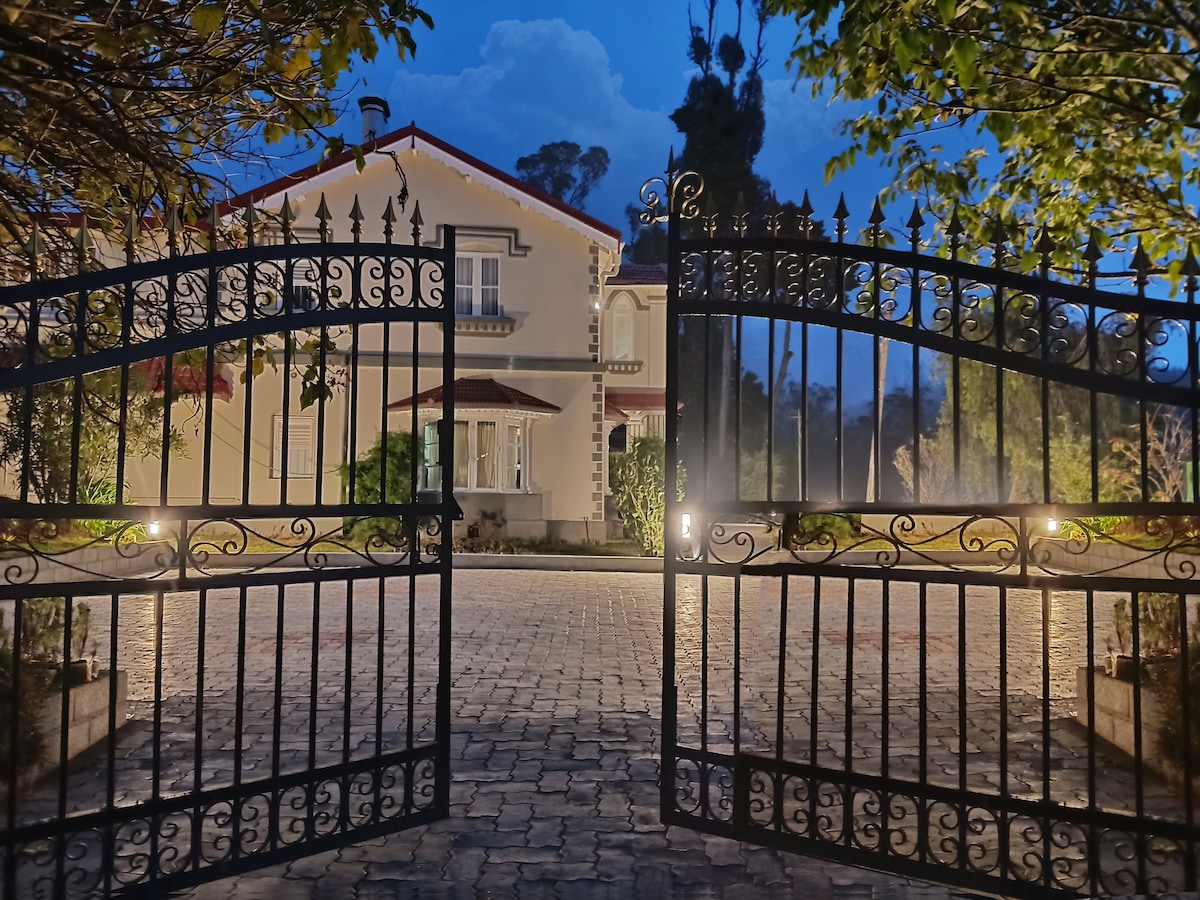 An ornate wrought iron gate opens to reveal a spacious brick pathway leading toward a charming two-story house. Soft lighting illuminates the pathway, framed by lush greenery and trees, creating a serene evening atmosphere.