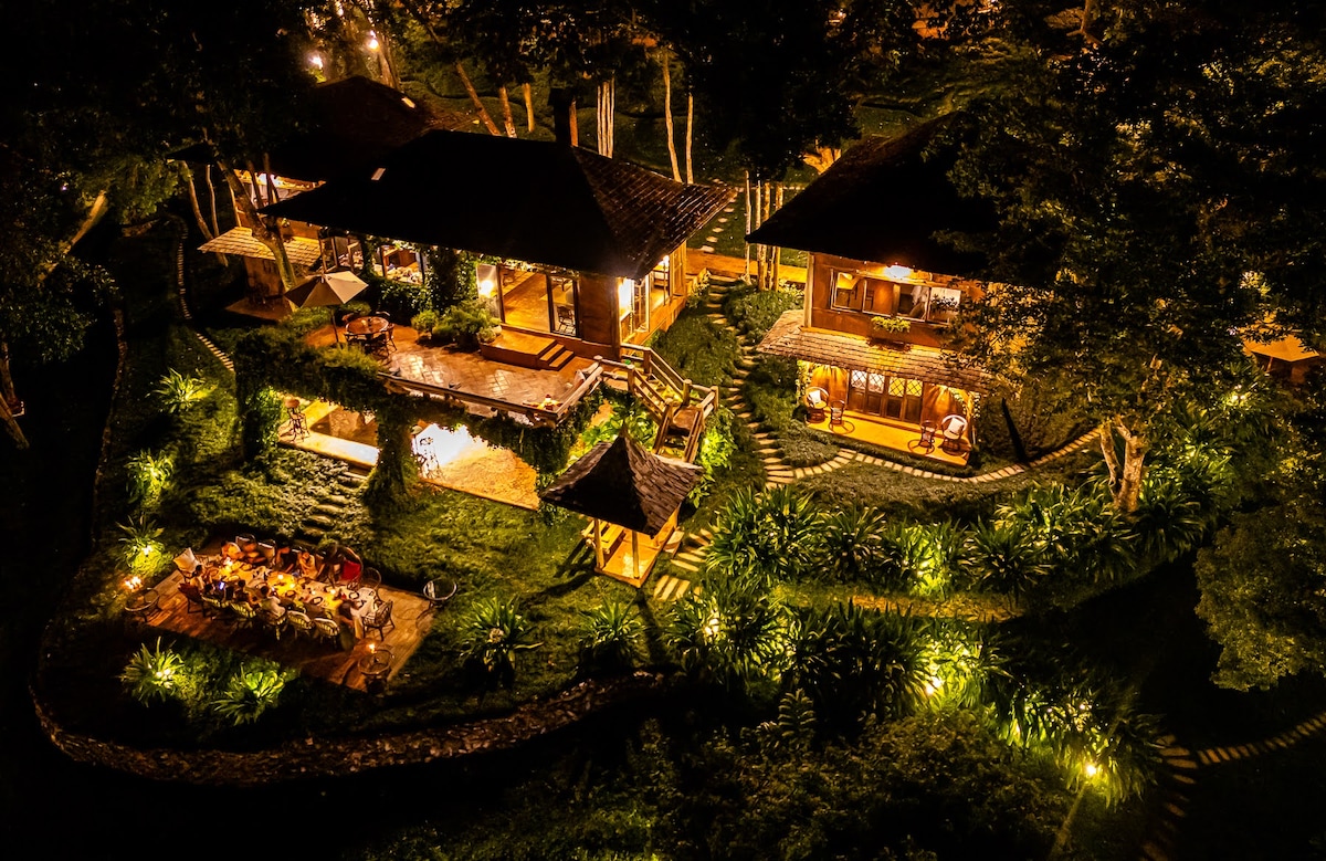 An aerial view captures two main buildings surrounded by lush greenery at night, softly illuminated by warm lights. A rustic outdoor dining area is set amidst the vibrant landscape, while pathways lead to a pool and cozy seating areas, enhancing the serene ambiance of the property.