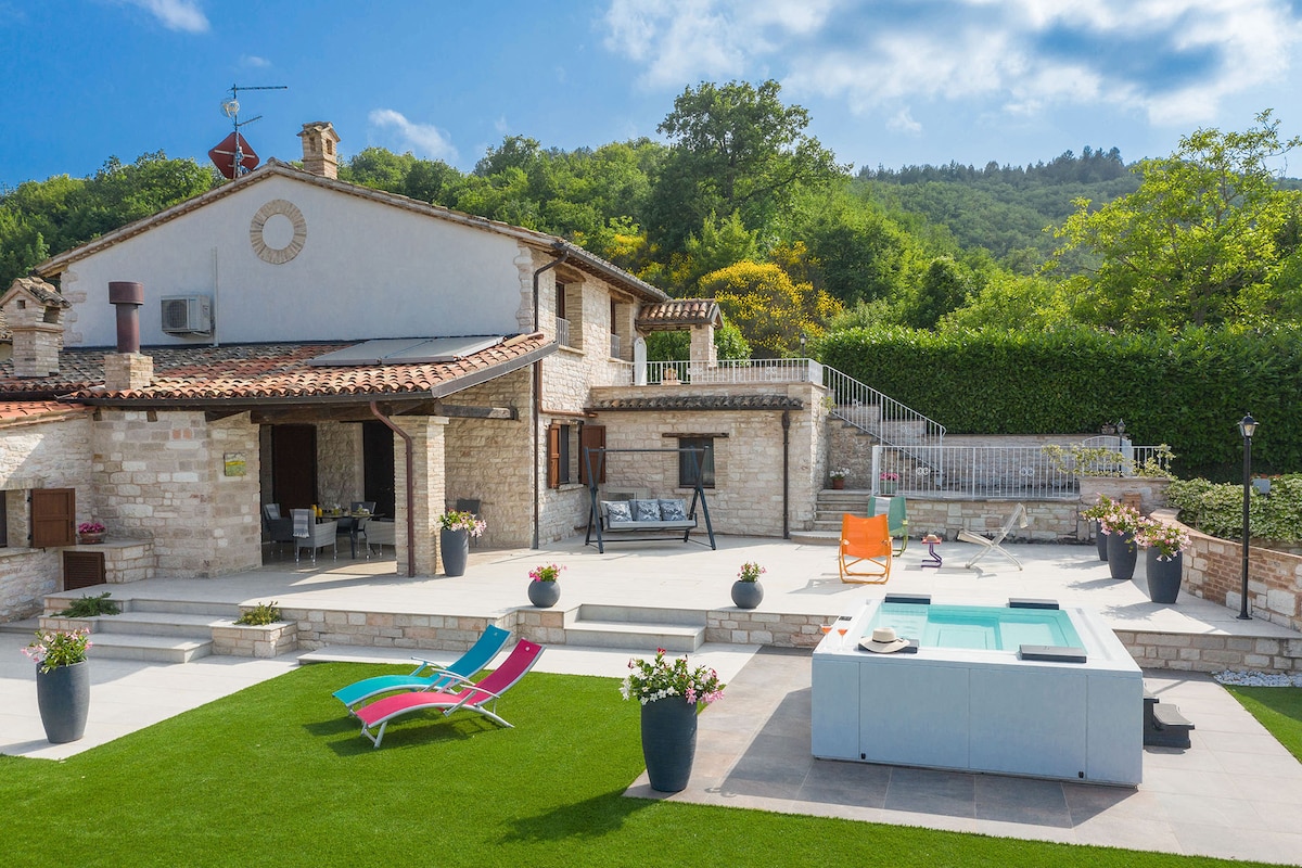 The outdoor area features a spacious terrace with a heated whirlpool tub, colorful sun loungers, and well-maintained synthetic grass. Flower pots with vibrant blooms are placed around the setting, enhancing the inviting atmosphere. A charming stone building provides a rustic backdrop against the lush green hillside.