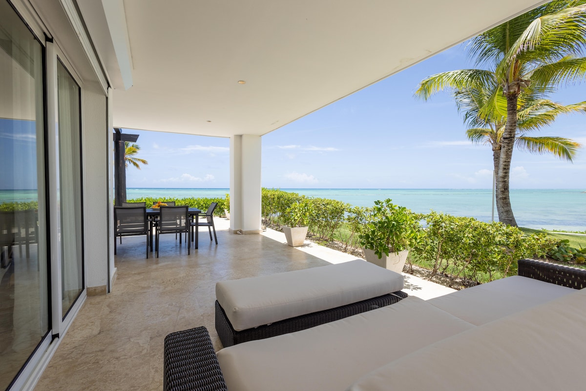 A spacious terrace overlooks the ocean, featuring a cozy seating area with lounge chairs. Surrounding greenery and planters enhance the outdoor setting, while a dining table for six is visible in the background. The tranquil sea and clear sky create a serene backdrop.