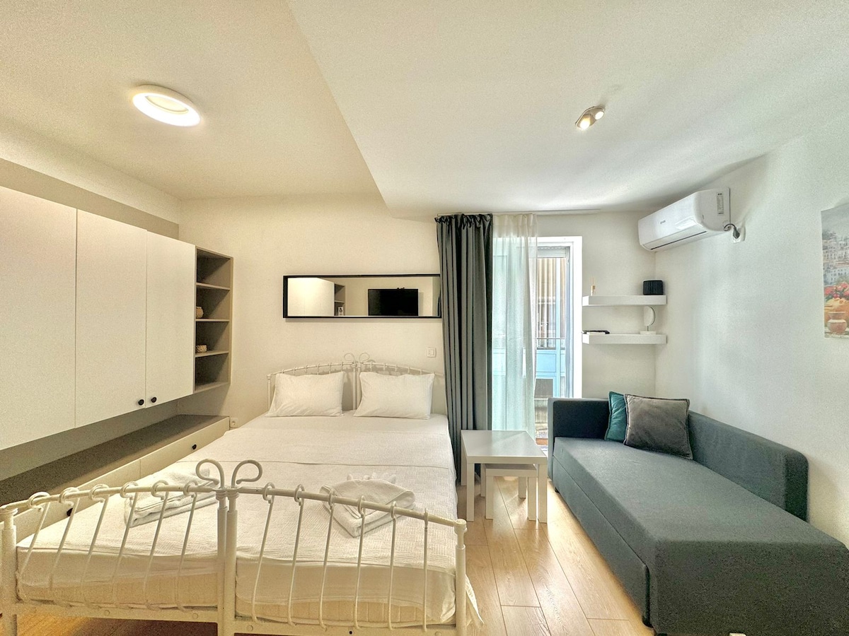 A studio apartment is presented with a spacious bed and a cozy seating area, complemented by a small table. The light wood flooring enhances the modern decor, while large windows provide natural light. A balcony is visible, adding to the overall charm of the space.