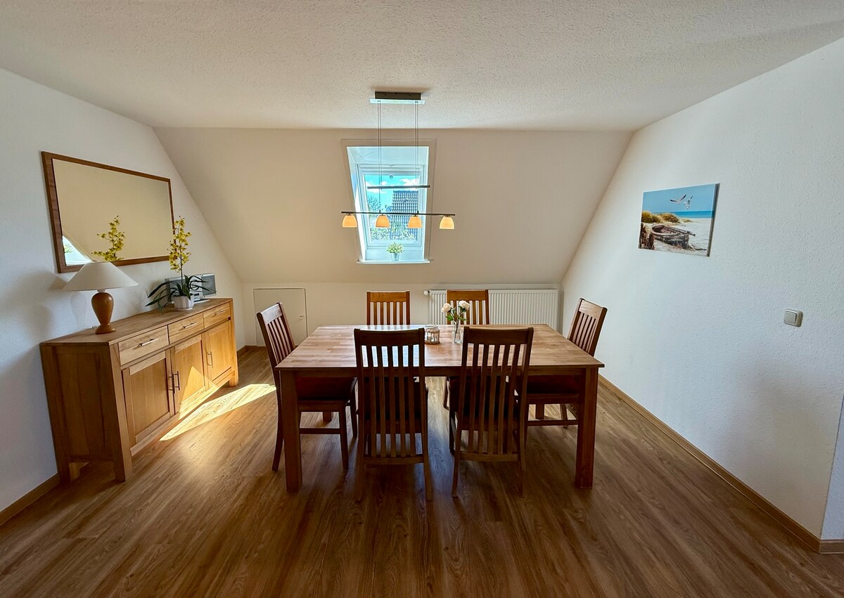A dining area features a large wooden table surrounded by six chairs, providing ample space for gatherings. Natural light enters through a skylight, illuminating the room. A sideboard and a mirror add functional decor, while a painting of a beach scene enhances the ambiance.