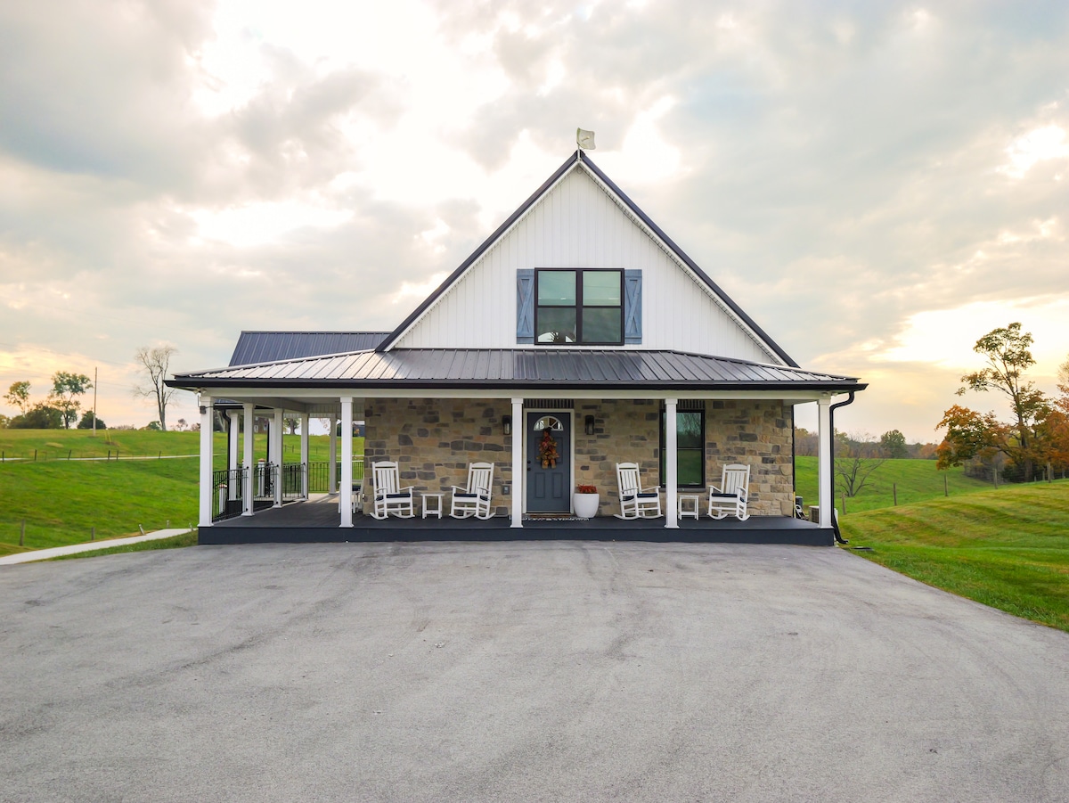 A charming cabin features a front porch adorned with several rocking chairs. The exterior combines stone and white siding, with large windows inviting natural light. Surrounding grassy fields are visible, under a cloudy sky that adds a serene backdrop.