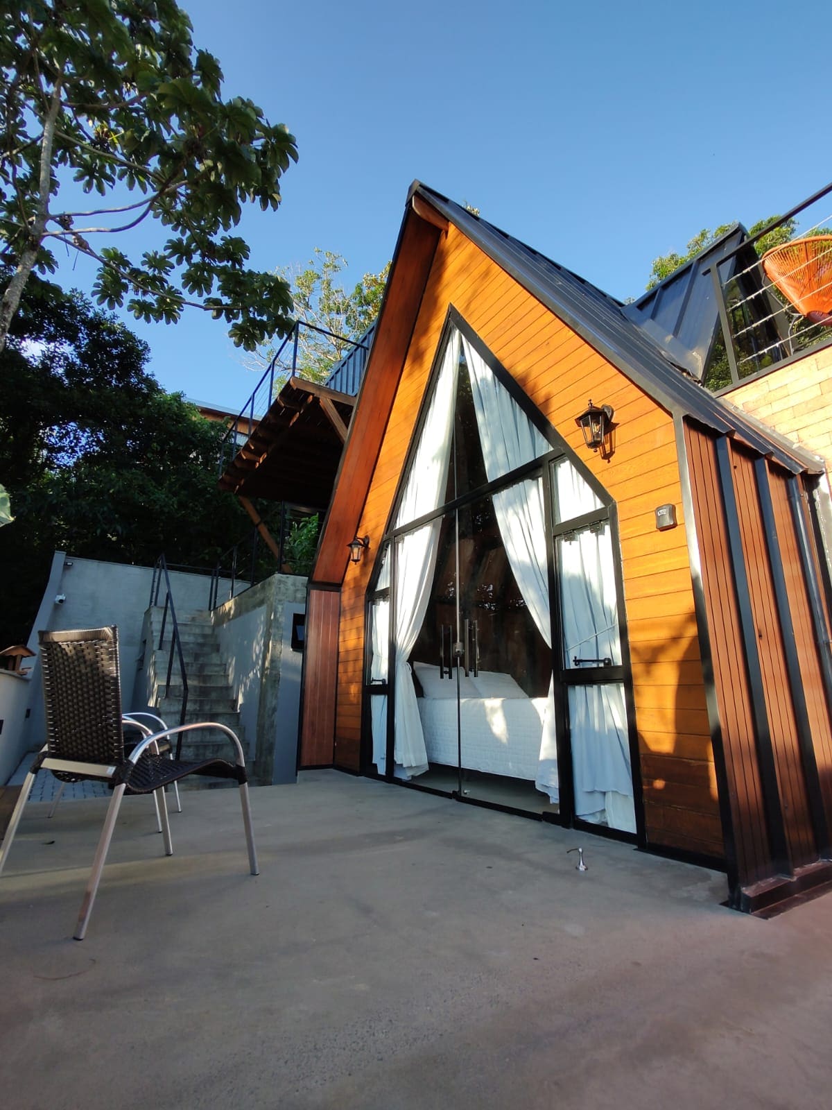 A charming chalet with an A-frame design is depicted, featuring large glass doors framed by white curtains. The wooden exterior reflects natural tones, while a small patio chair is positioned nearby. Lush greenery surrounds the building, adding to the serene environment.