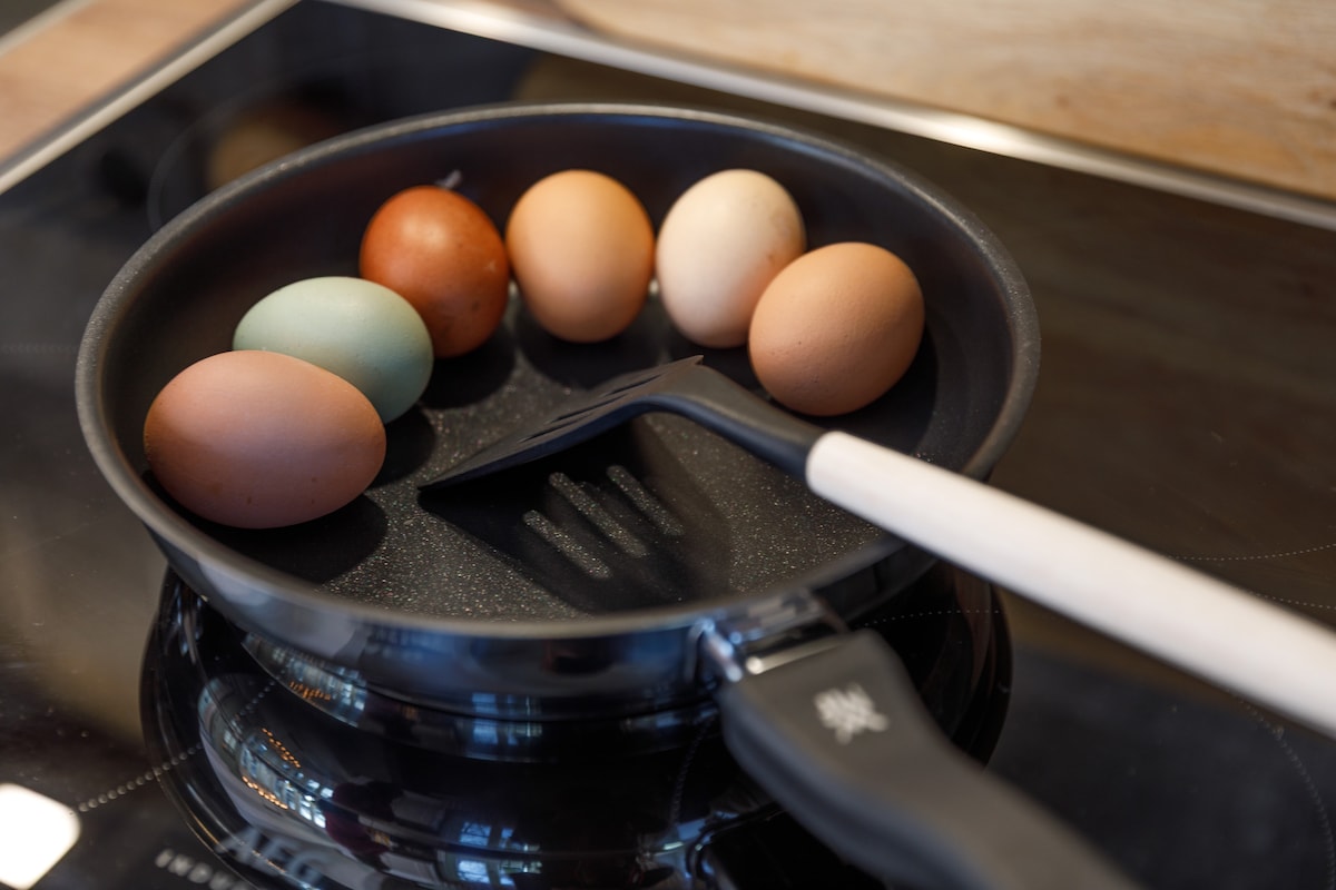 A frying pan is positioned on a stovetop, containing a selection of seven eggs in various colors, including brown, cream, and blue. A spatula with a white handle rests beside the pan, indicating preparation for cooking.