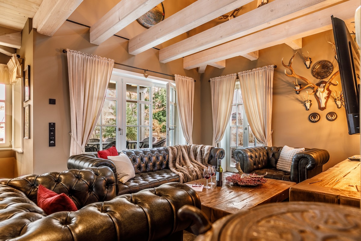 A cozy living area features large leather sofas arranged around a rustic wooden coffee table. Natural light streams through expansive glass doors, enhancing the warm tones of the walls. Elegant curtains frame the windows, while decorative items, including wall-mounted antlers, add character to the space.