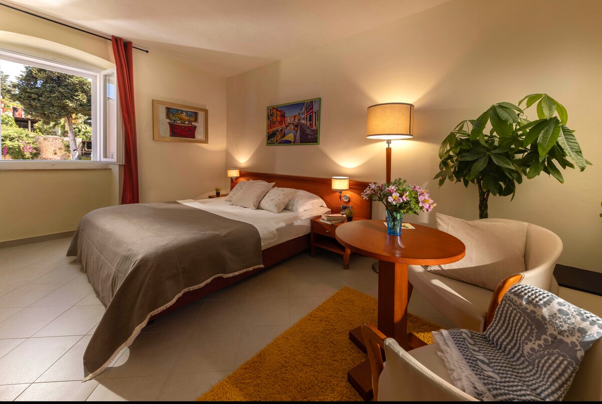 A cozy double room features a comfortable bed, complemented by soft bedding. A small wooden table with a vase of flowers and a chair is present. Natural light enters through a window adorned with red curtains, highlighting the neutral color palette of the room.