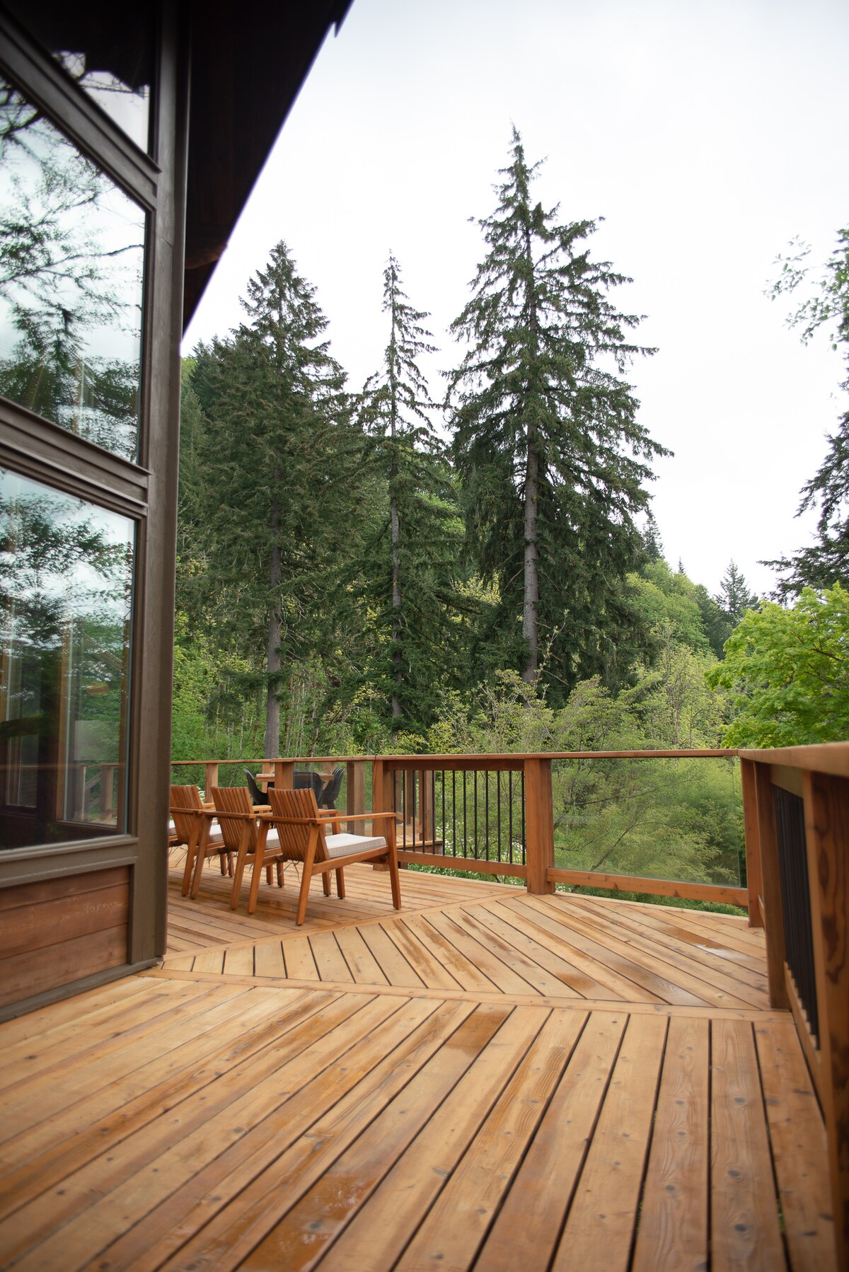 A spacious wooden deck provides seating with chairs overlooking a lush landscape of tall evergreen trees. The structure features large windows reflecting the surrounding greenery, integrating the indoor and outdoor environments.