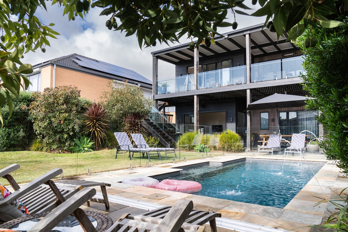 The outdoor space features a private, solar-heated in-ground pool surrounded by lounge chairs. A multi-level home sits in the background, with an expansive deck showcasing shaded seating. Lush landscaping creates a serene atmosphere, complemented by gentle poolside splashes.