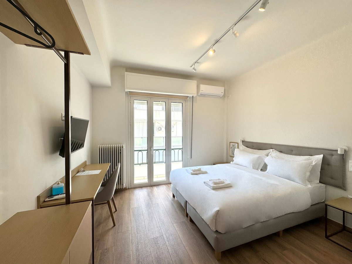 A well-lit guest room features a spacious bed with neatly arranged linens and towels. A desk and chair are positioned beside a wall-mounted television. Large glass doors lead to a balcony, allowing natural light to fill the space, while a minimalistic design enhances the room's modern aesthetic.