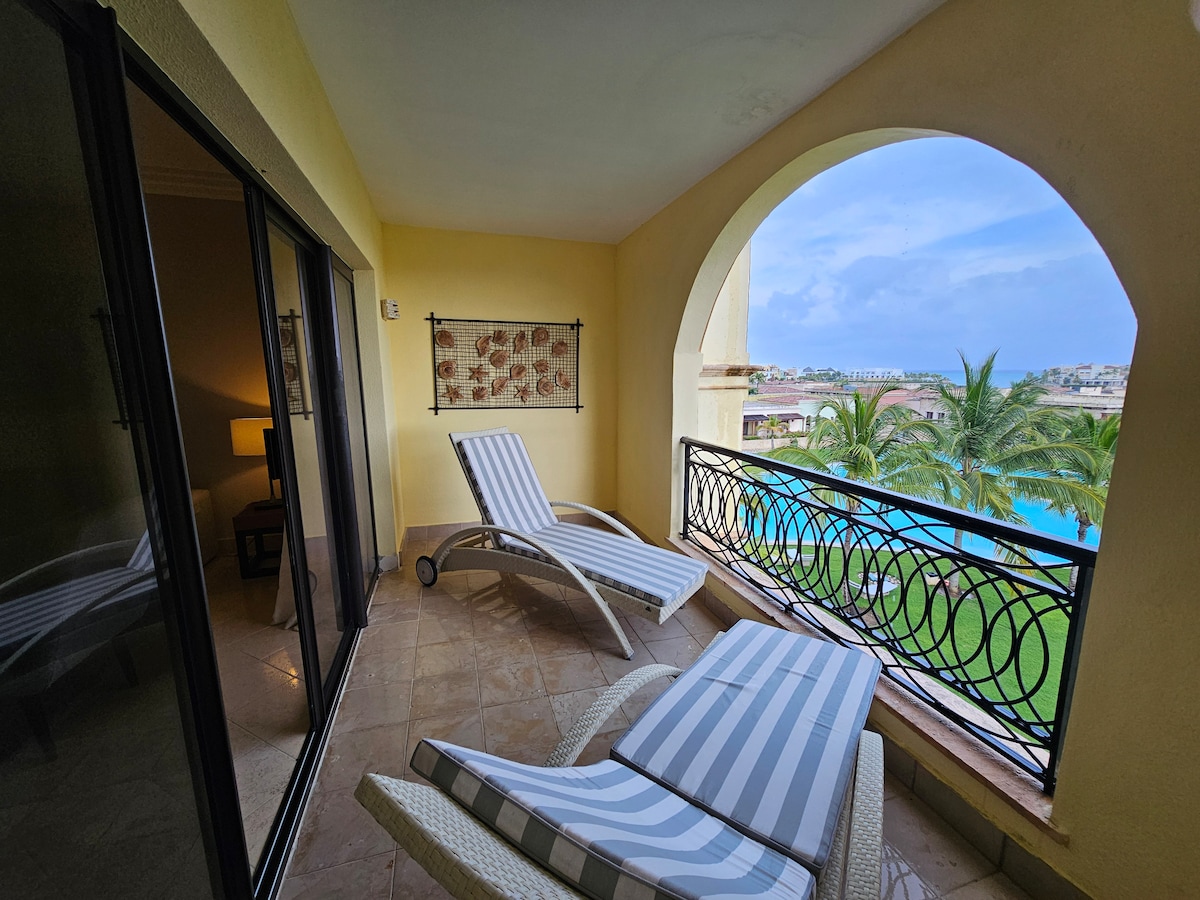A sunlit balcony features two comfortable loungers with striped cushions. An arched entrance frames a view of lush palm trees and a serene blue pool. The neutral-toned walls and tiled flooring lend a calm ambiance to the outdoor space.