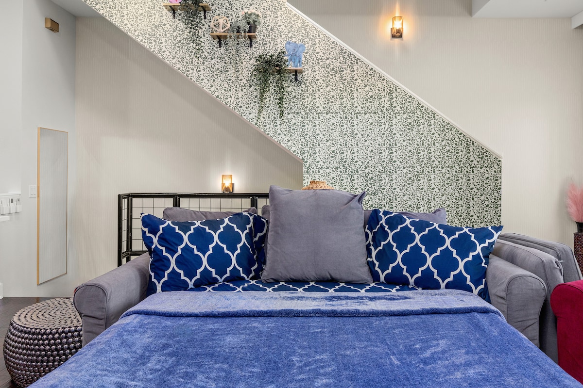 A cozy living area features a spacious sofa adorned with blue and white patterned cushions. A soft blue throw blanket adds comfort. The backdrop showcases a staircase with a decorative wall, enhanced by greenery and subtle lighting fixtures.