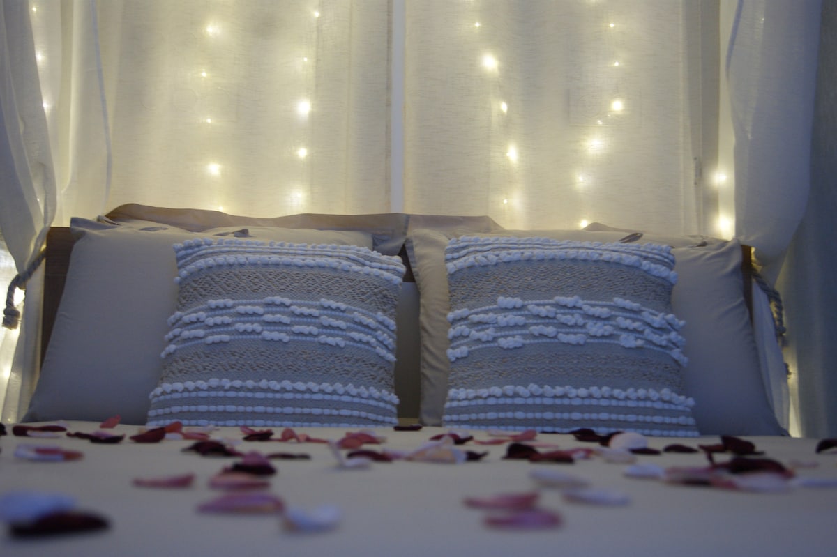 A cozy bed is adorned with decorative pillows, featuring textured designs. Soft, pastel-hued rose petals are scattered across the bedspread, adding a romantic touch. Behind the bed, twinkling string lights cast a gentle glow, creating a serene and inviting ambiance.