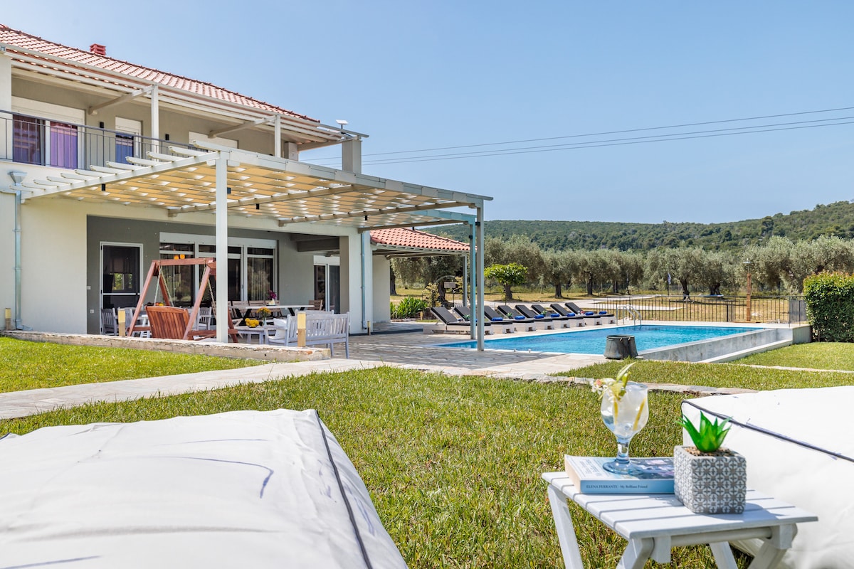 An outdoor patio area is visible, featuring a shaded seating space with a modern swing and lounge chairs. A refreshing pool is positioned in the background, surrounded by a lush green lawn that leads to an olive grove, providing a peaceful setting.