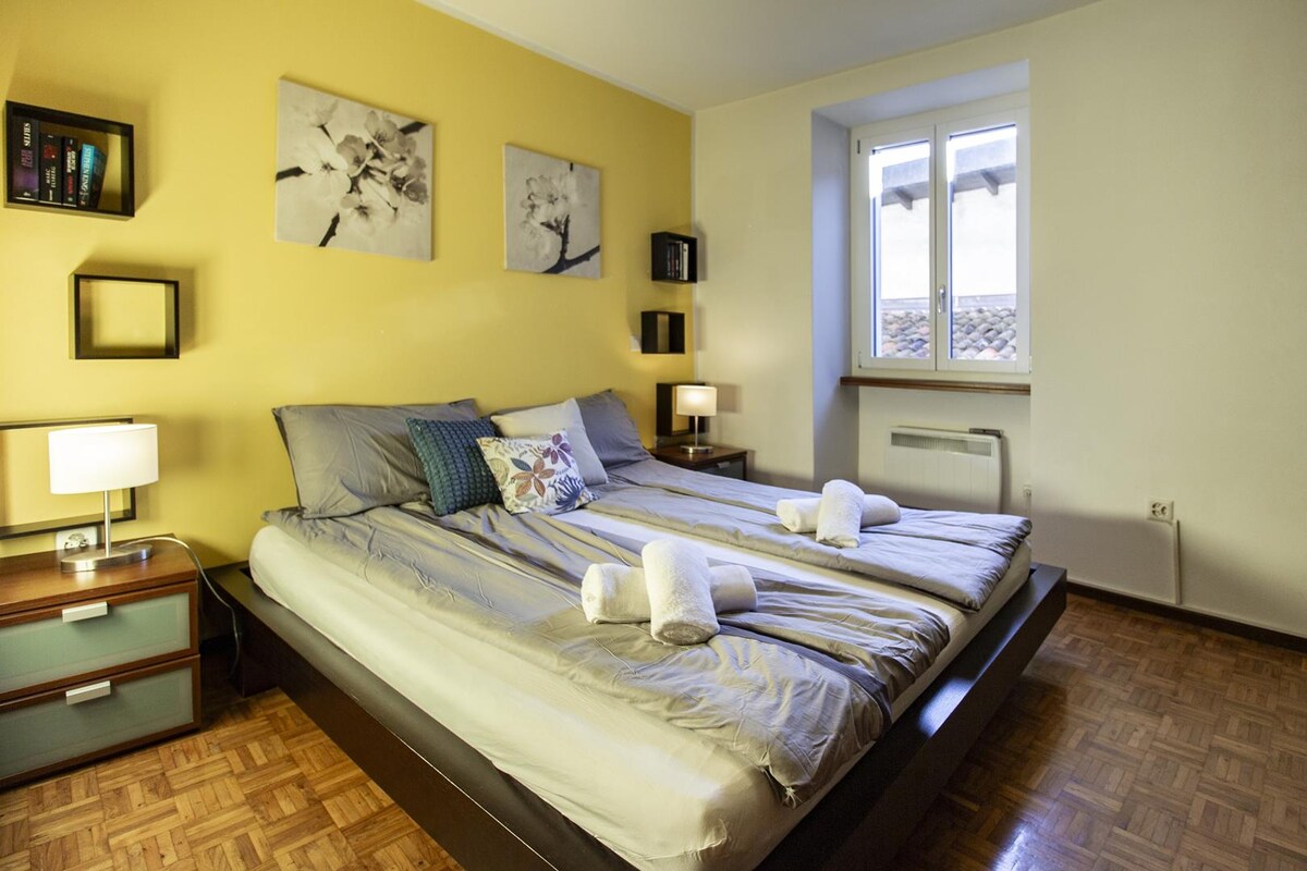 ห้องสว่างสดใสพร้อมเตียงคู่หมอนตกแต่งและผ้าขนหนูพับ ผนังสีเหลืองภาพวาดดอกไม้และโต๊ะข้างเตียงพร้อมโคมไฟสร้างบรรยากาศที่อบอุ่น