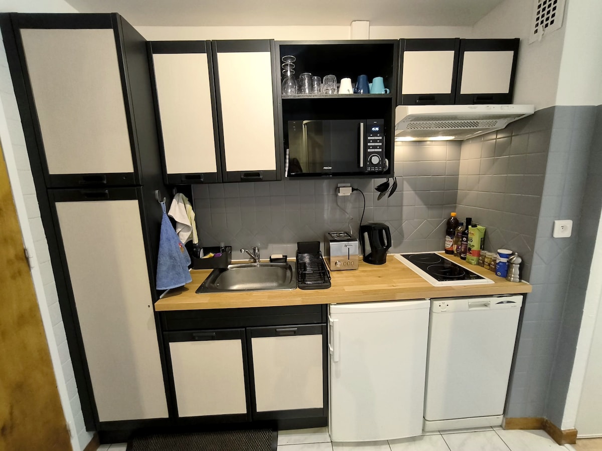 The kitchen is compact and equipped with essential appliances including a microwave, kettle, and stovetop. Ample cabinetry provides storage, while a sink is located under a wall-mounted shelf. Counter space is complemented by a light wood surface, contributing to a functional layout.