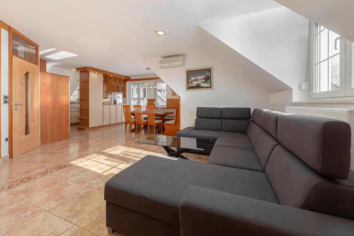 An open living space is showcased, featuring a comfortable dark sectional sofa positioned next to a sleek black coffee table. Natural light floods the area through large windows, illuminating the modern dining table set for six. A kitchen is visible in the background, enhancing the space's functionality.