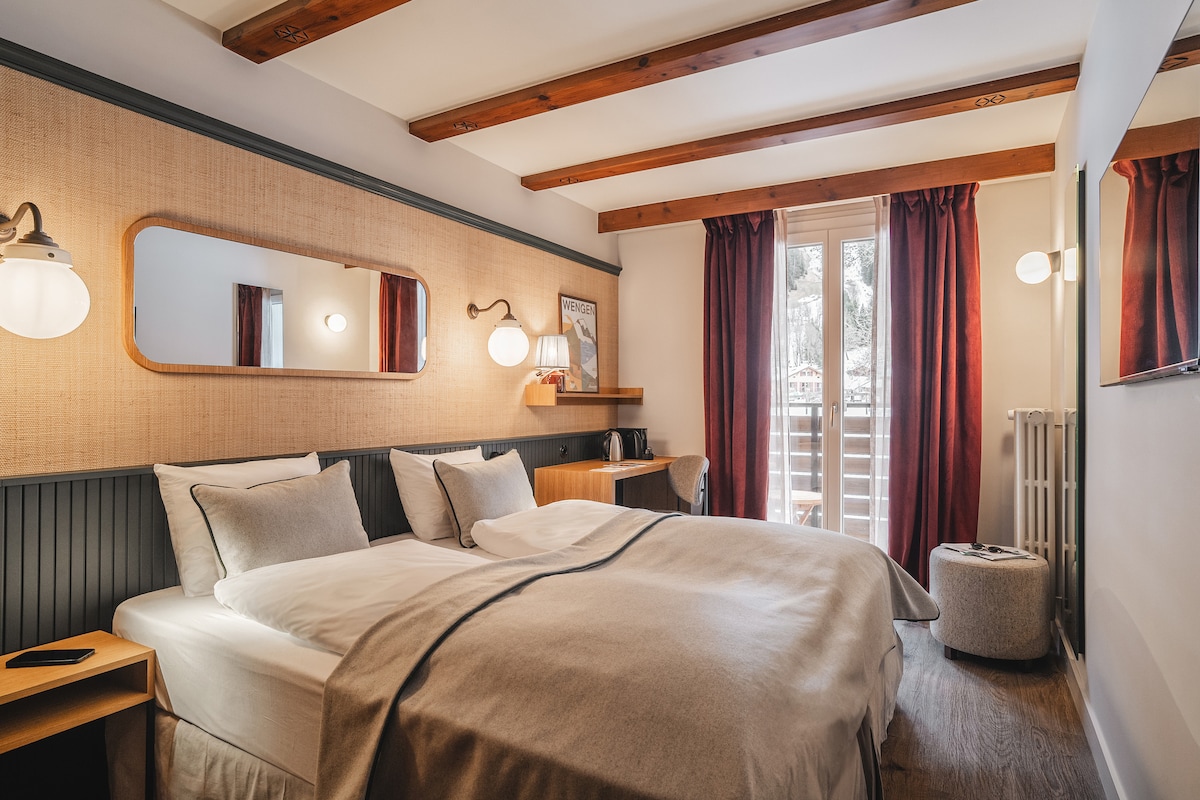 A welcoming bedroom is furnished with a spacious bed featuring light linens and numerous pillows. Natural light enters through a window with dark red curtains, and a wooden shelf holds a kettle and cups. A large mirror and decorative lighting enhance the room's comfort.