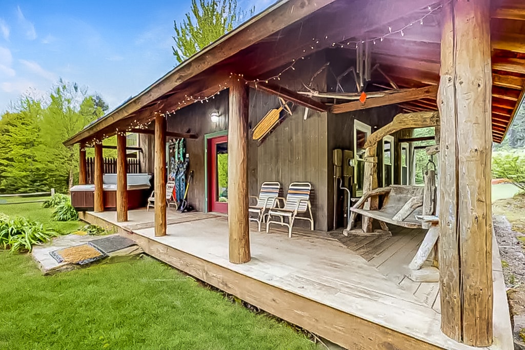 A spacious wraparound deck, highlighted by natural wooden beams, features multiple seating areas and a private hot tub. Surrounded by lush greenery, the inviting outdoor space is enhanced with string lights, providing a perfect setting for relaxation and enjoyment.