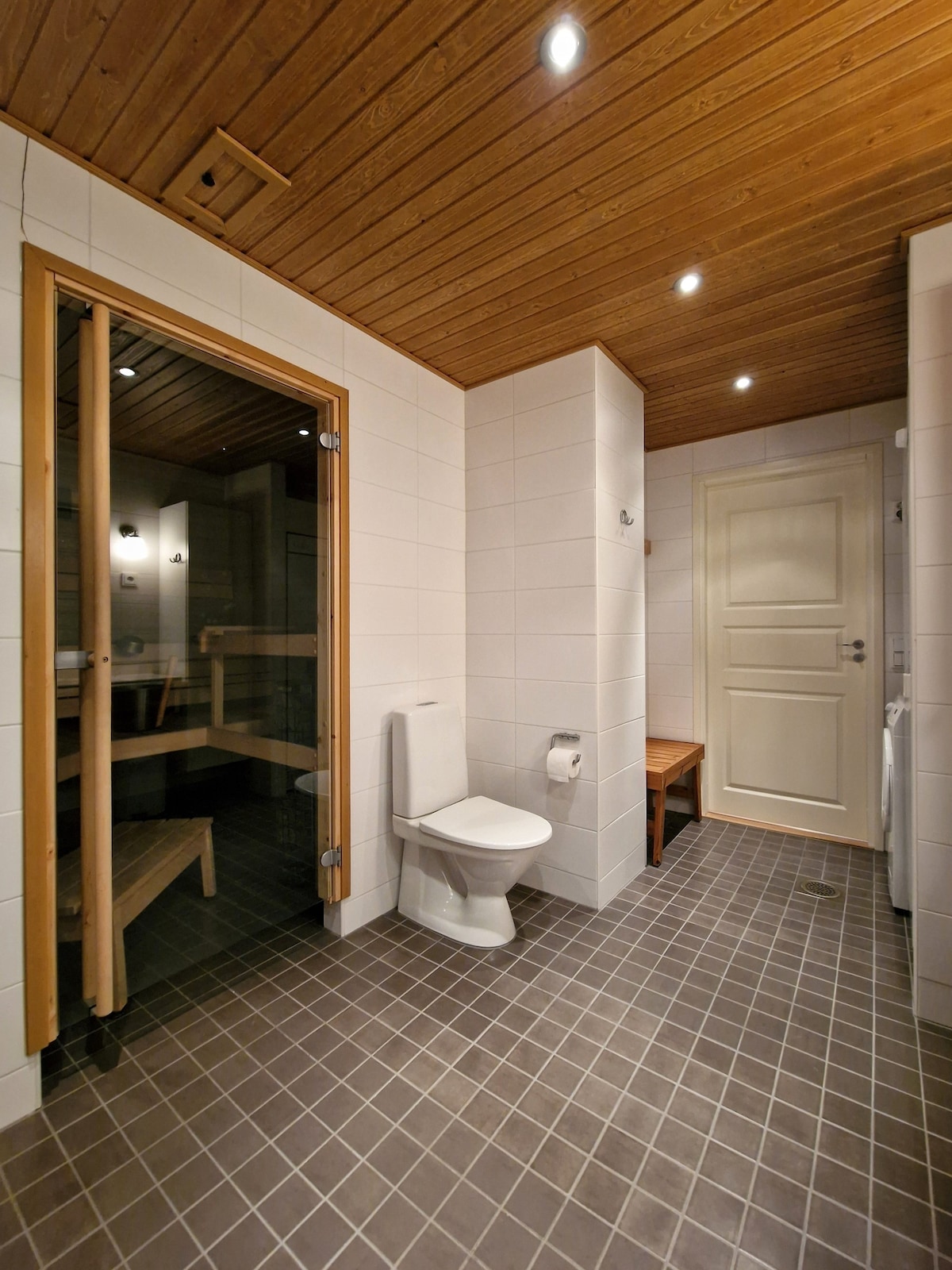 A modern bathroom is shown featuring a wooden sauna area with benches and glass doors. A toilet is positioned near the entrance, with a wooden bench on one side. The walls are tiled in white and gray, complemented by warm wood accents in the ceiling.