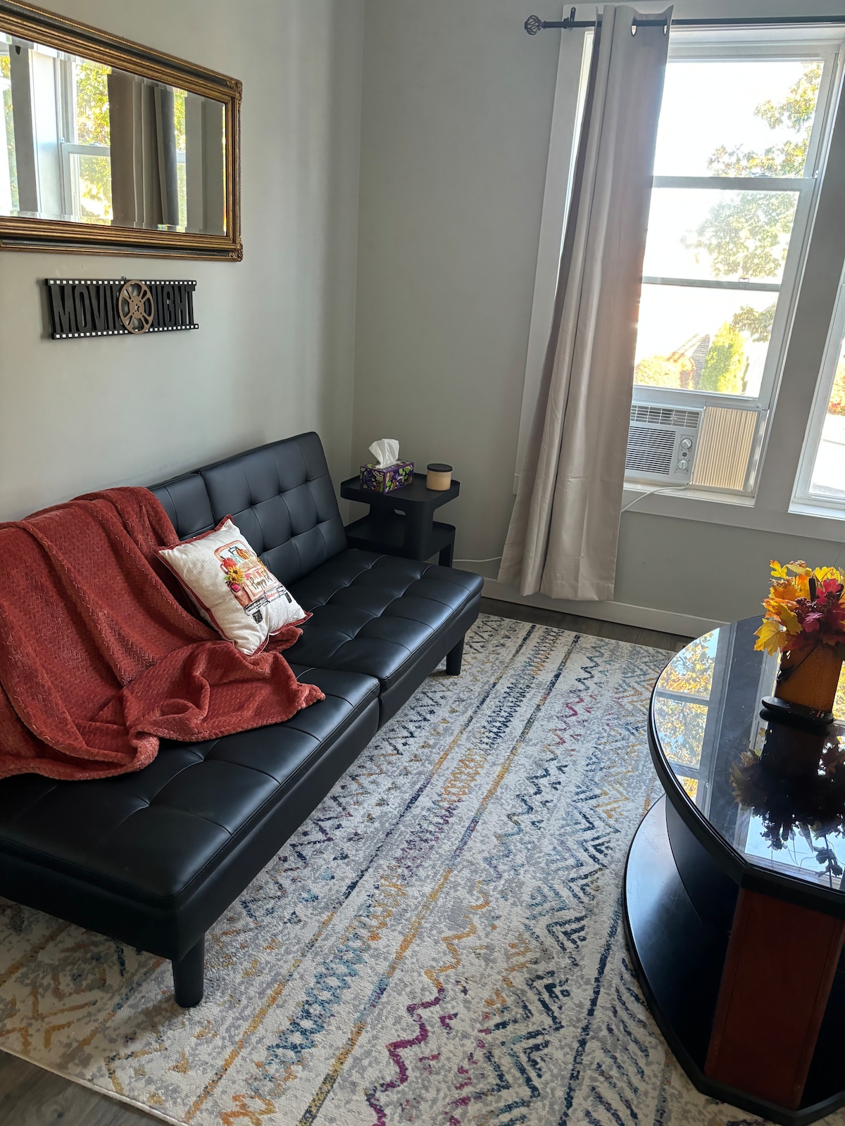 A modern seating area features a sleek black futon adorned with a decorative pillow and a cozy red throw. A round table with a small vase of flowers complements the space, while natural light streams in through large windows draped with light curtains.
