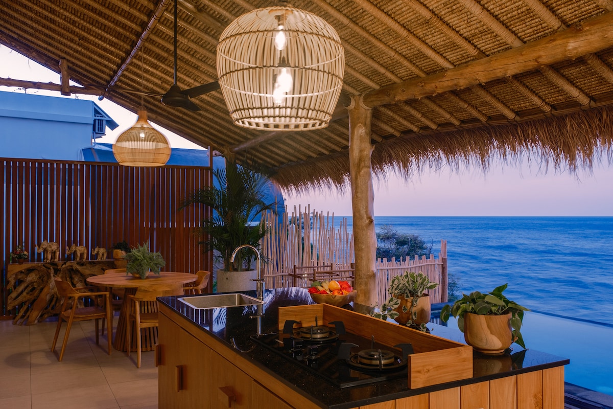 An open-concept kitchenette features a black countertop with a gas stove and a modern wooden design. Natural elements, including potted plants, enhance the space. A dining area with wooden chairs is positioned nearby, and expansive ocean views can be seen through the open layout.