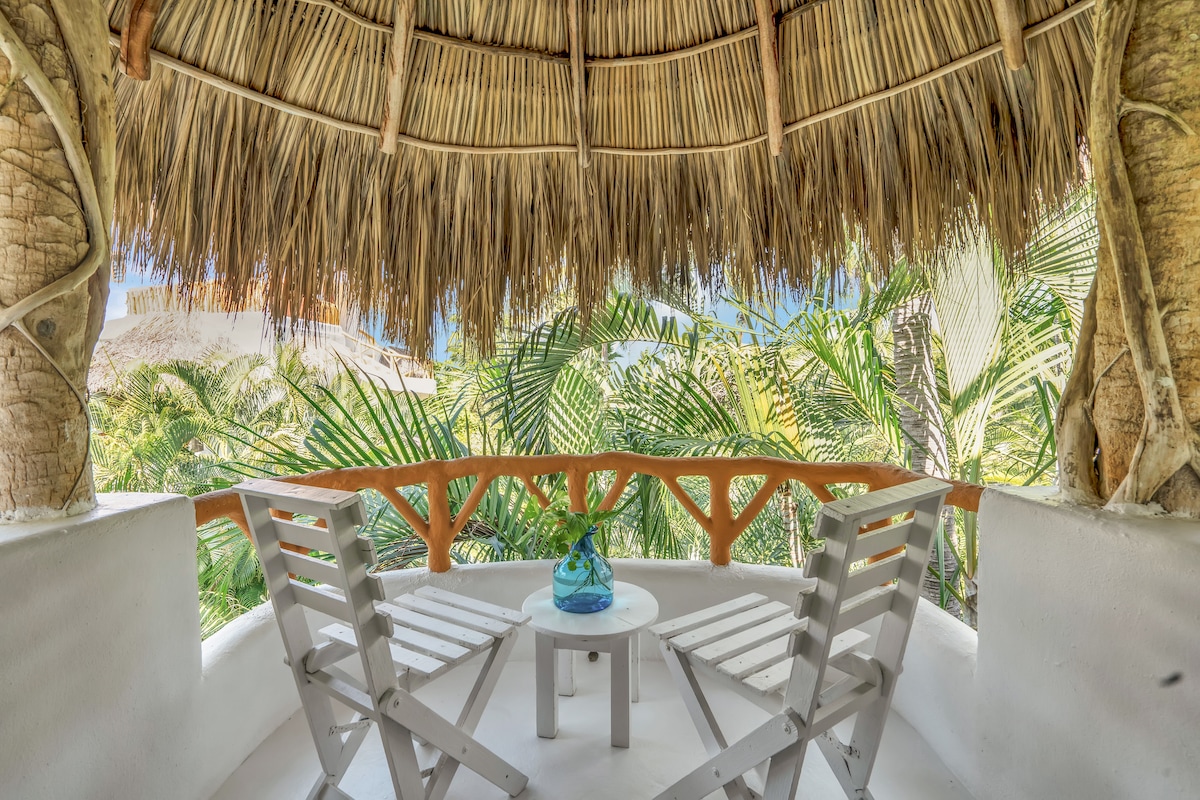 A covered terrace is highlighted by a thatched roof, featuring two white chairs and a small round table. Lush greenery is visible through the railings, creating a serene and private atmosphere. Natural textures and colors enhance the connection to the surrounding tropical environment.