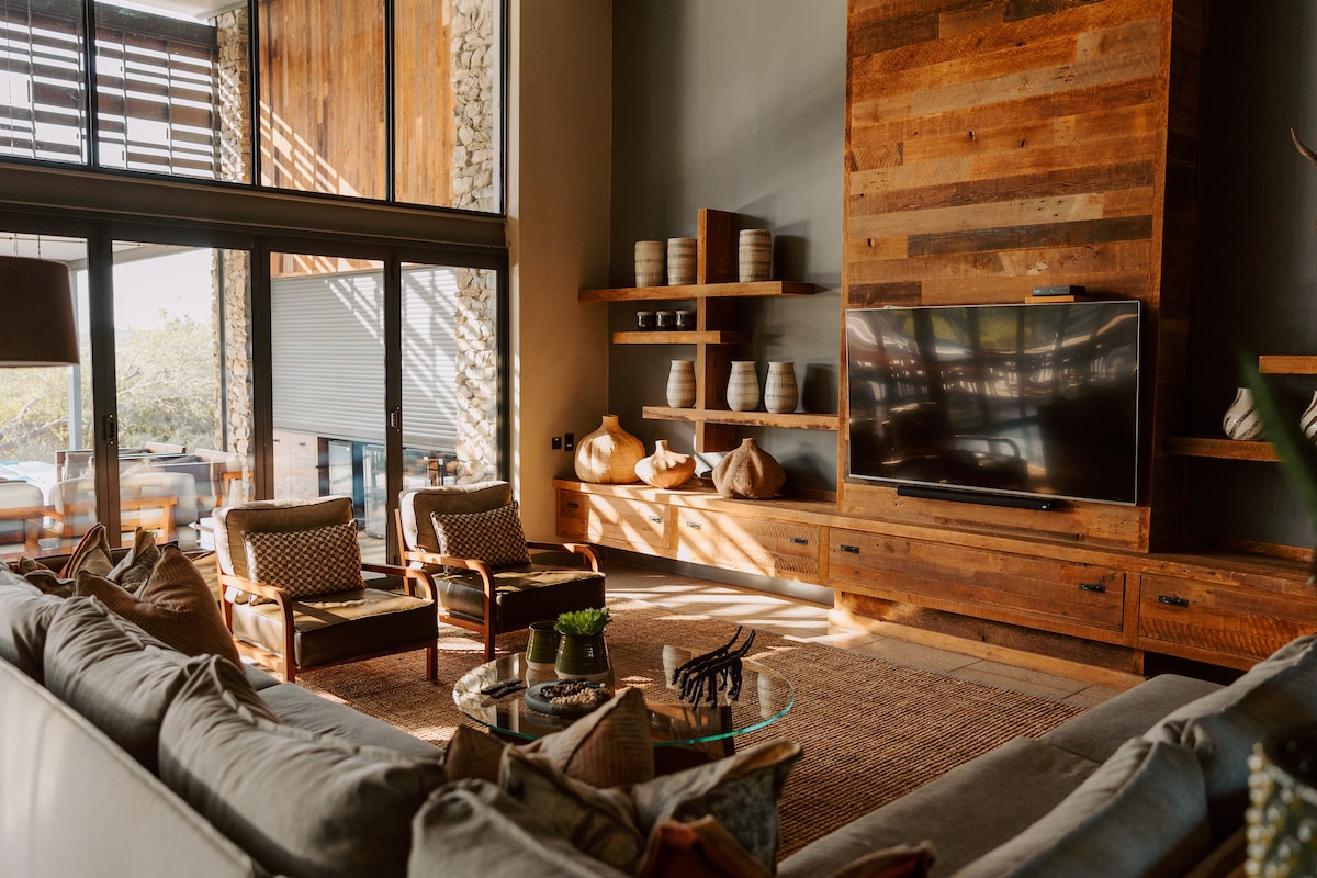 A spacious living area features a large, comfortable sofa with textured cushions, complemented by stylish armchairs. A wooden media unit is equipped with a flat-screen TV, while decorative pottery is displayed on the shelves. Natural light filters in through expansive glass doors, illuminating the warm wood accents.