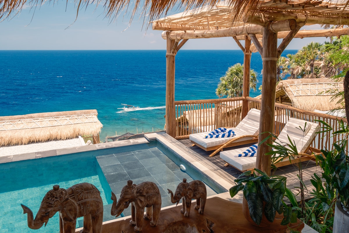 An inviting infinity pool is visible, surrounded by sun loungers with blue and white striped cushions. In the foreground, wooden elephant sculptures are displayed on a surface, while the expansive ocean view provides a backdrop under clear skies.