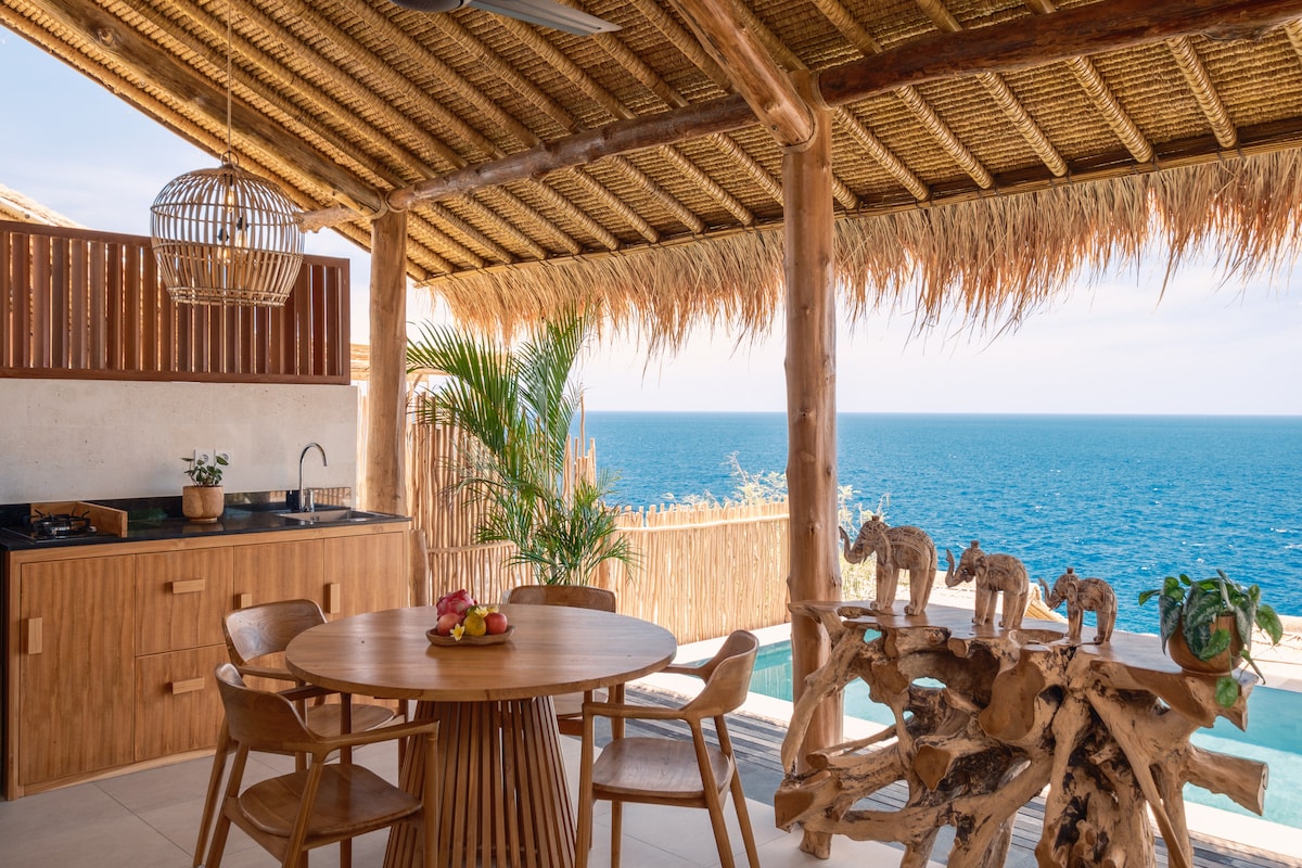 An open-concept terrace is seen, featuring a round wooden dining table with four chairs. A kitchenette with bamboo accents is positioned in the background, while the turquoise sea spans the horizon, framed by natural materials and coastal decor.