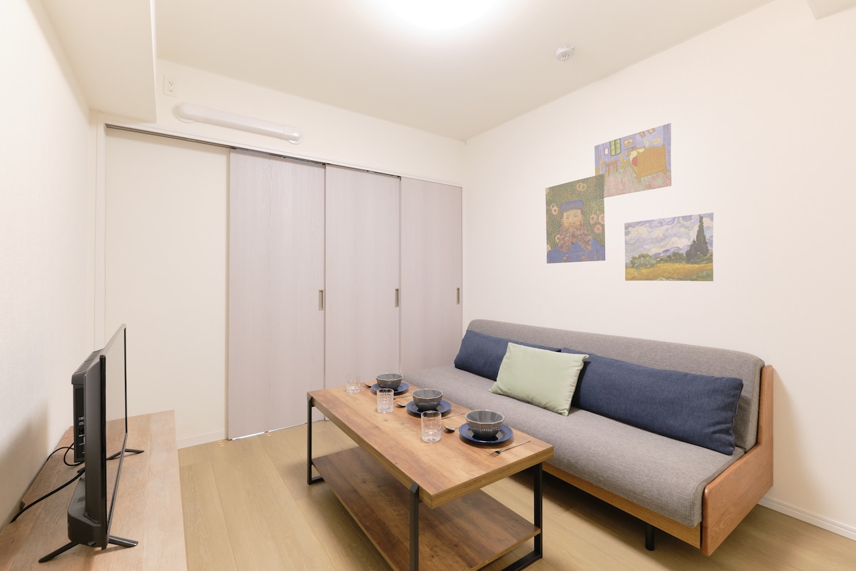 A modern living area features a comfortable sofa bed and a wooden coffee table set with dishes and glasses. Two artworks adorn the wall, while a television sits on a wooden console. Sliding doors lead to additional room space.