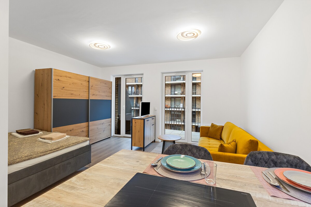 A modern living space features a wooden dining table set with colorful plates and napkins. A comfortable yellow sofa is positioned nearby, complemented by a stylish bed with neatly arranged textiles. Large windows allow natural light, while built-in shelves display neatly organized items.
