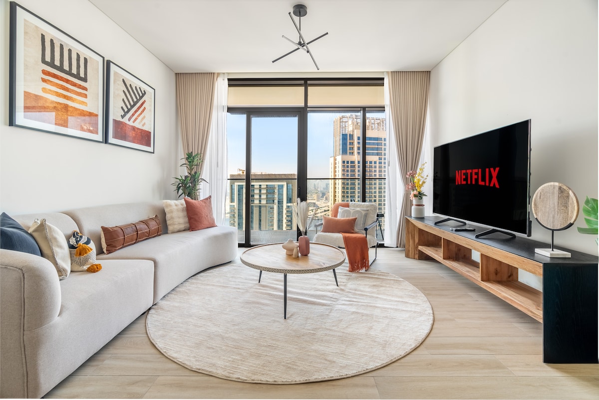 A bright living room features floor-to-ceiling windows offering views of the city. A light-colored, curved sofa is accented with decorative cushions. A sleek round coffee table is positioned in front of a large flat-screen TV. A small plant and artwork adorn the walls.