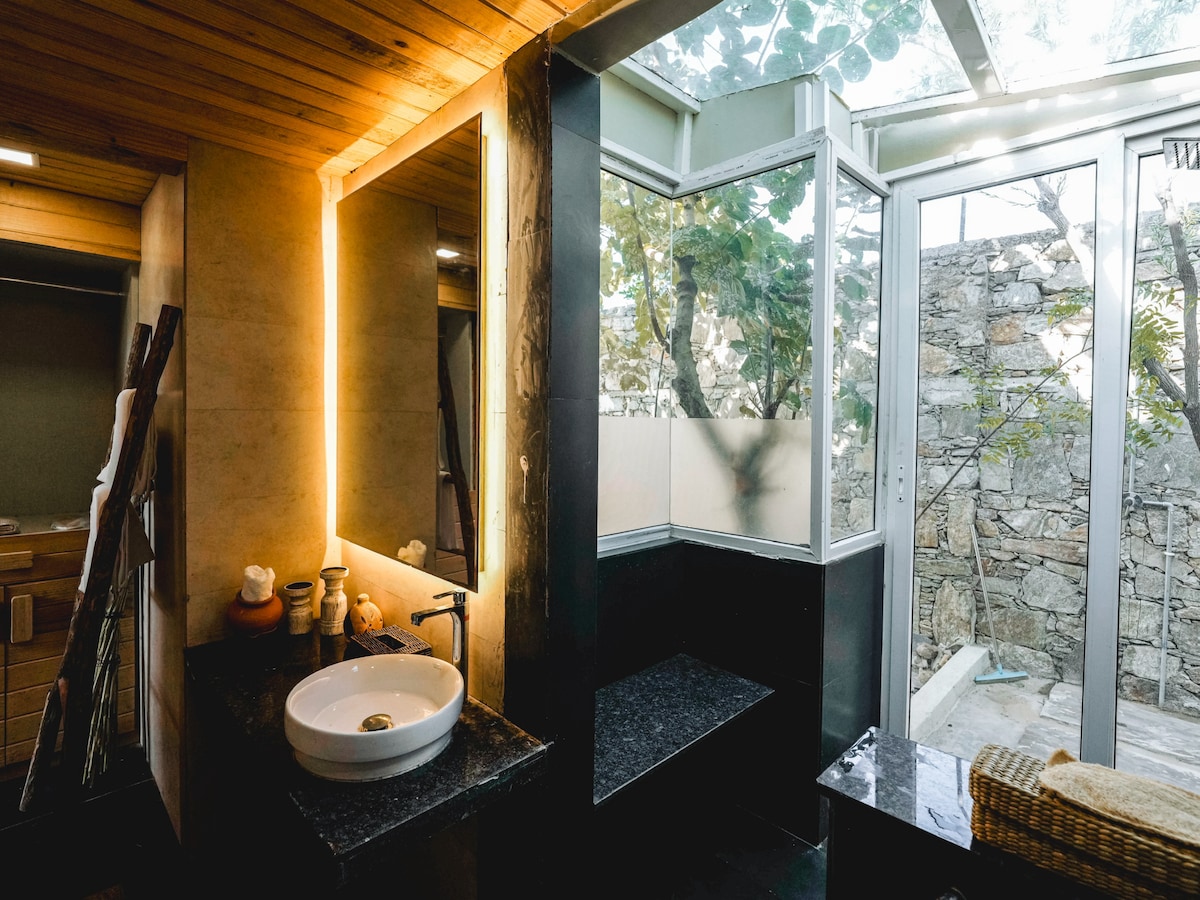 A modern bathroom features a circular sink on a dark countertop, complemented by a large mirror illuminated by ambient lighting. Floor-to-ceiling windows allow natural light to fill the space, offering views of greenery, while a stone wall adds an organic touch.