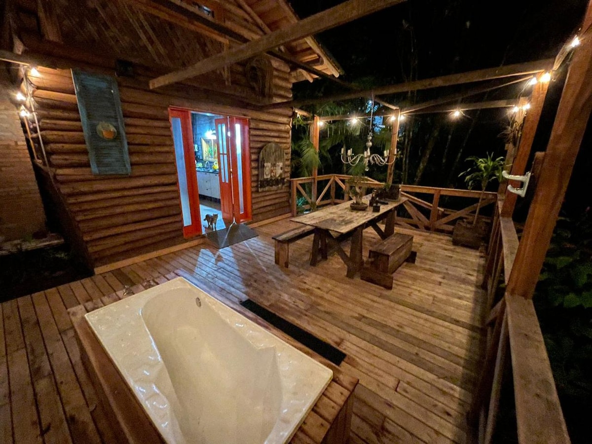 An outdoor space features a wooden deck illuminated by string lights, creating a cozy atmosphere. A white, freestanding bathtub sits beside a rustic wooden dining table, surrounded by natural greenery, providing a serene setting for relaxation and outdoor dining.