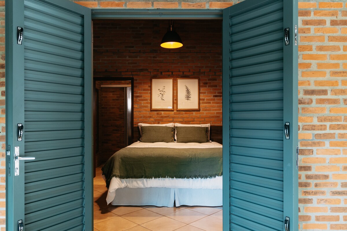 Double doors open to reveal a spacious bedroom featuring a large bed adorned with soft linens and pillows. Exposed brick walls give a rustic feel, while simple artwork adorns the walls, enhancing the room's natural ambiance. Warm lighting is provided by a pendant light.
