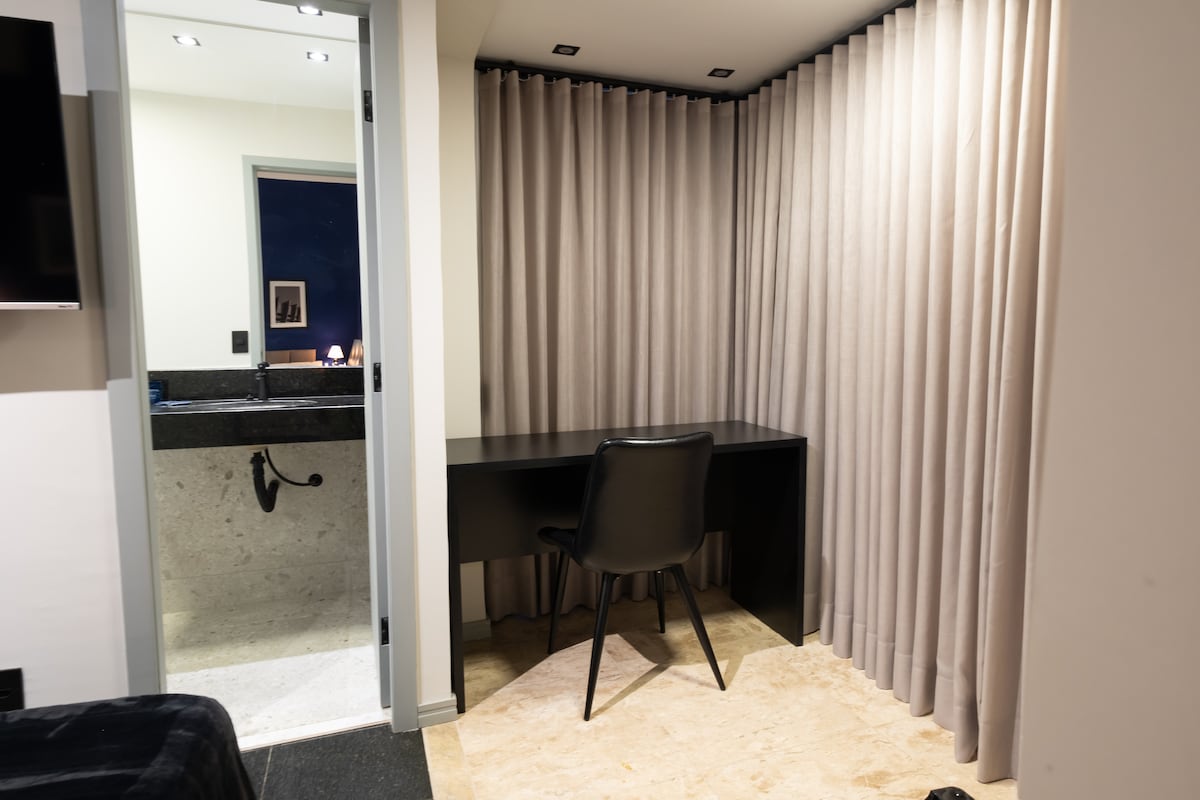 A dedicated workspace is featured within the room, including a sleek black desk and a comfortable chair. Soft curtains in neutral tones provide privacy, while a small sink area is visible in the background, emphasizing functionality within the space.