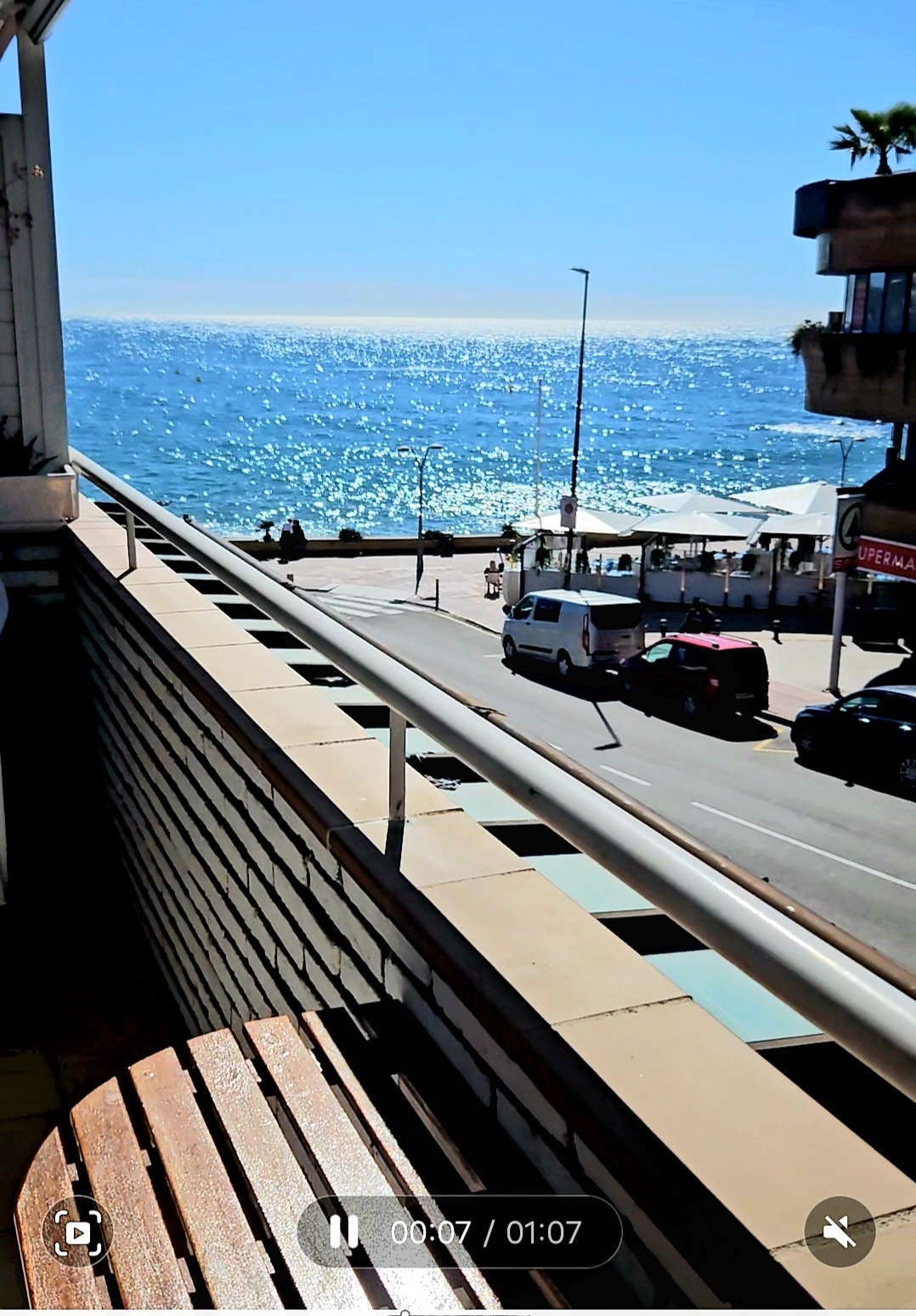 A balcony offers a clear view of the shimmering sea under a bright blue sky. The vibrant water reflects sunlight, and the coastline is visible in the distance. Prominent features include a wooden bench and a railing that adds to the open and breezy atmosphere.